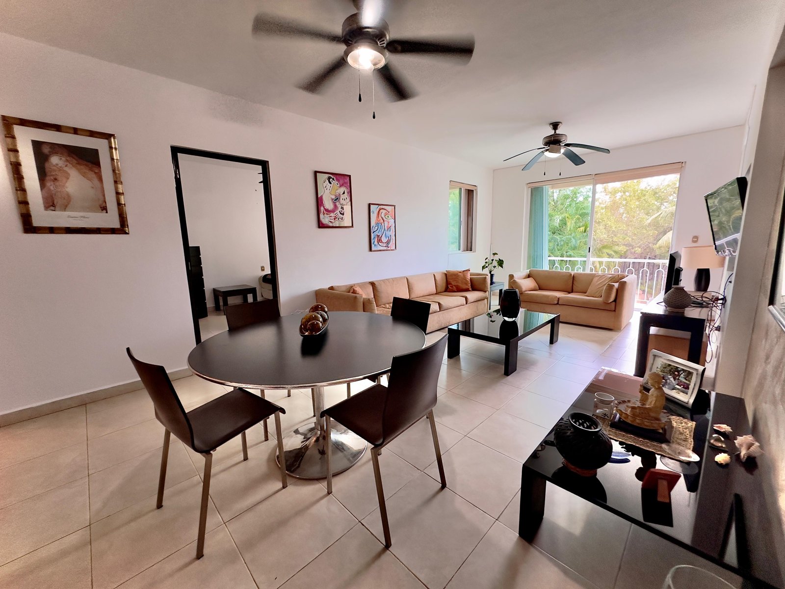 Sala-comedor integrada con dos ventiladores de techo, mesa redonda negra, sofás, puerta corrediza con vista a vegetación tropical