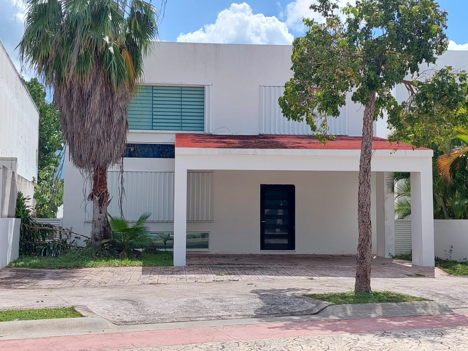 Fachada de dos plantas en blanco con detalle rojo, cochera techada, puerta negra y árboles.