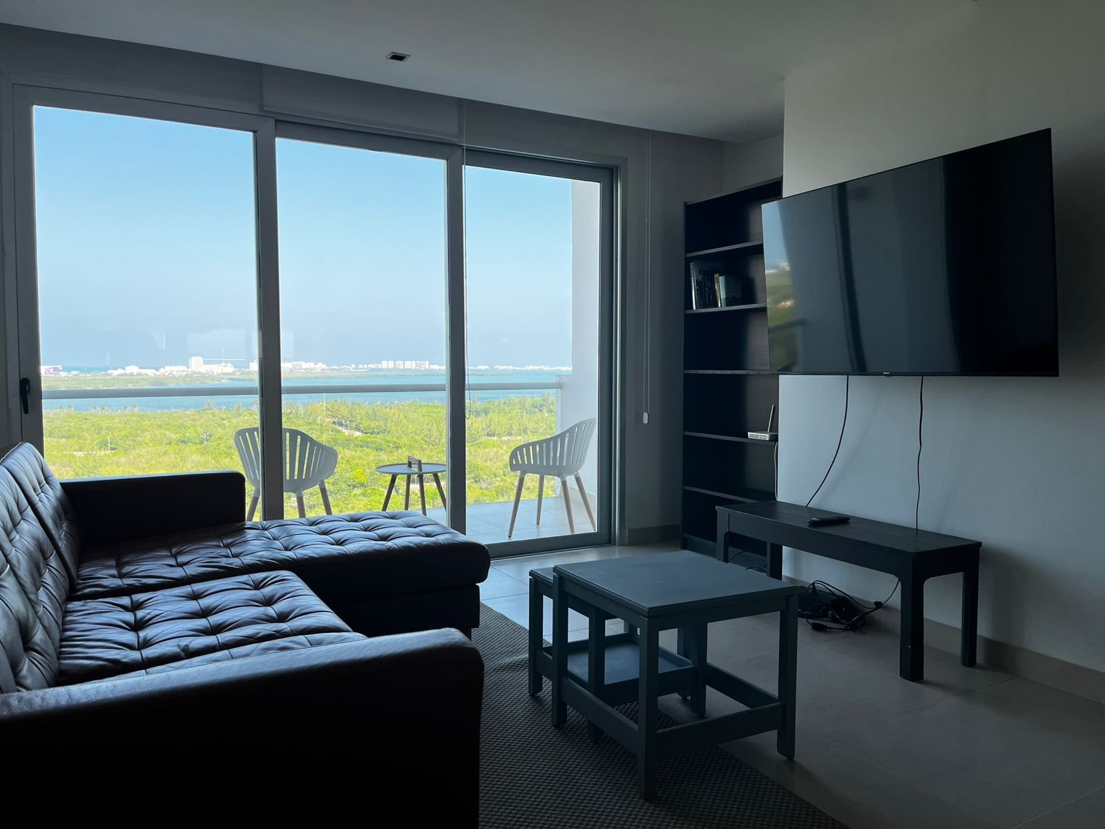 Sala con sofá seccional oscuro, puerta corrediza de piso a techo con vista a laguna Nichupté, manglares y cielo azul